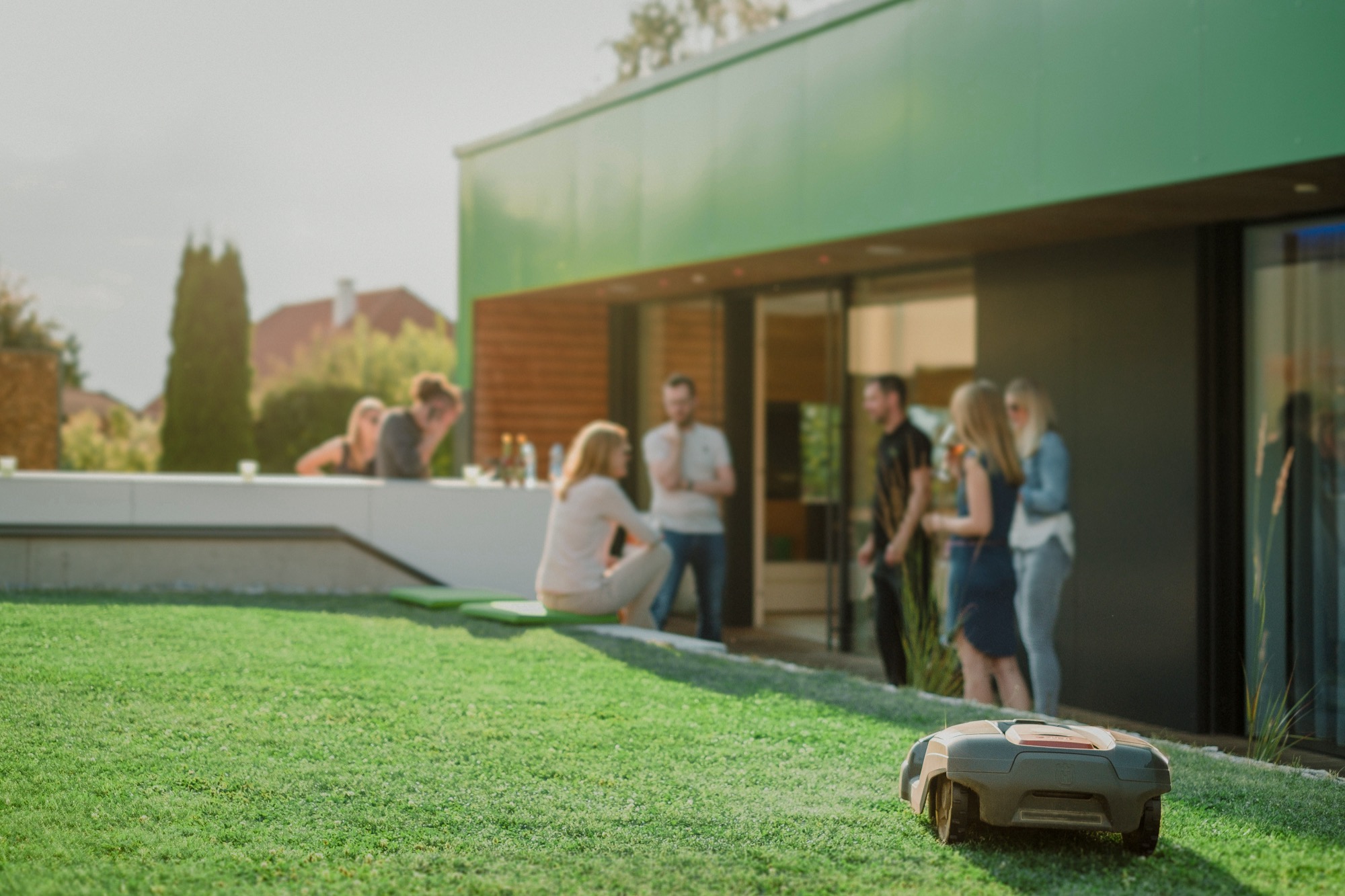 Friends gathered on a sunlit terrace while a robotic mower tends the lawn